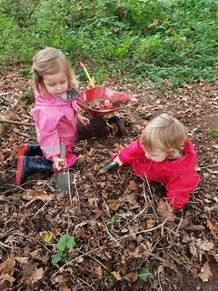 Two toddlers digging in leaf litter with trowels beside a wheelbarrow