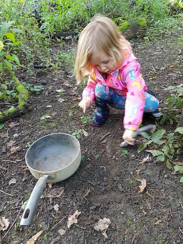 A child crouched on the forest floor, digging beside an old saucepan